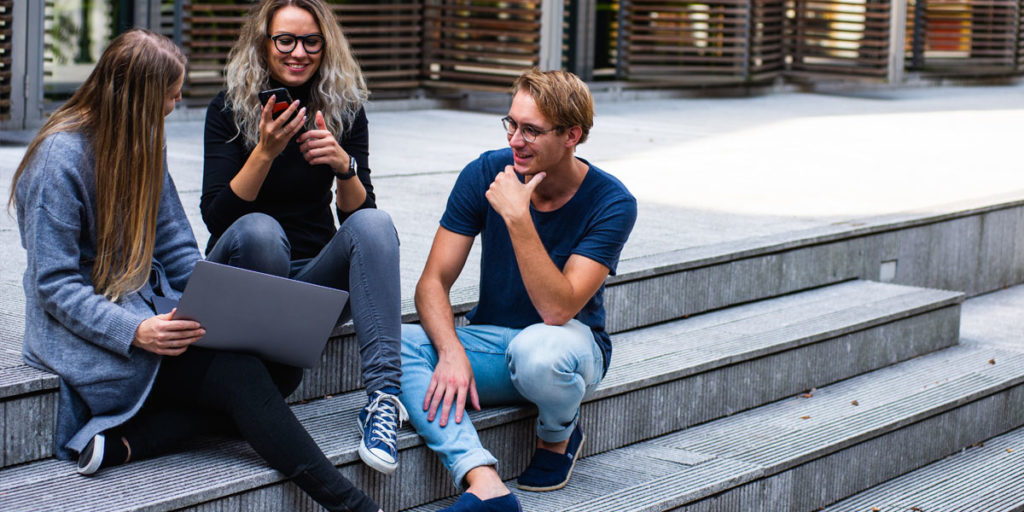 Group of young adults sitting on outdoor concrete steps looking at a laptop screen.
