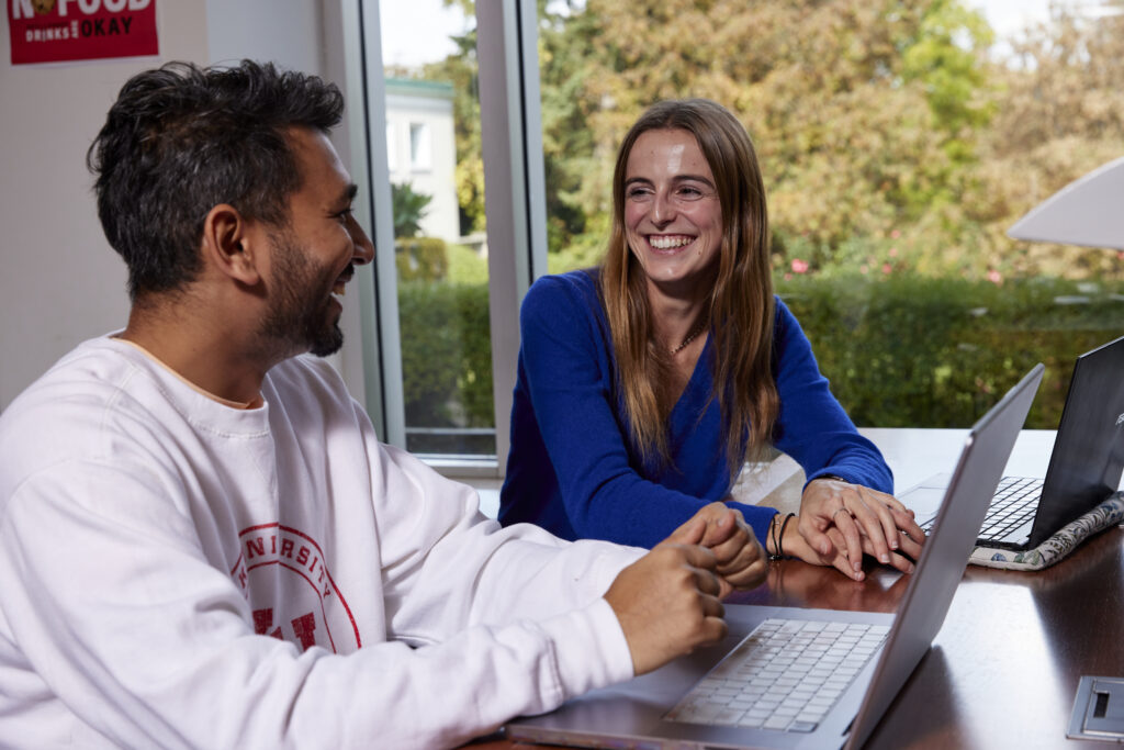 two students sitting at a desk with laptops open and in conversation