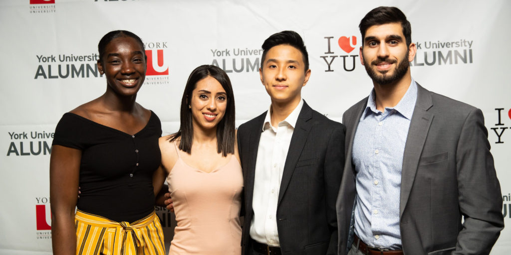 A group of young York University alumni pose in front of a York University alumni backdrop at an event