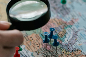 Hand holding a magnifying glass over a map with colored pins.